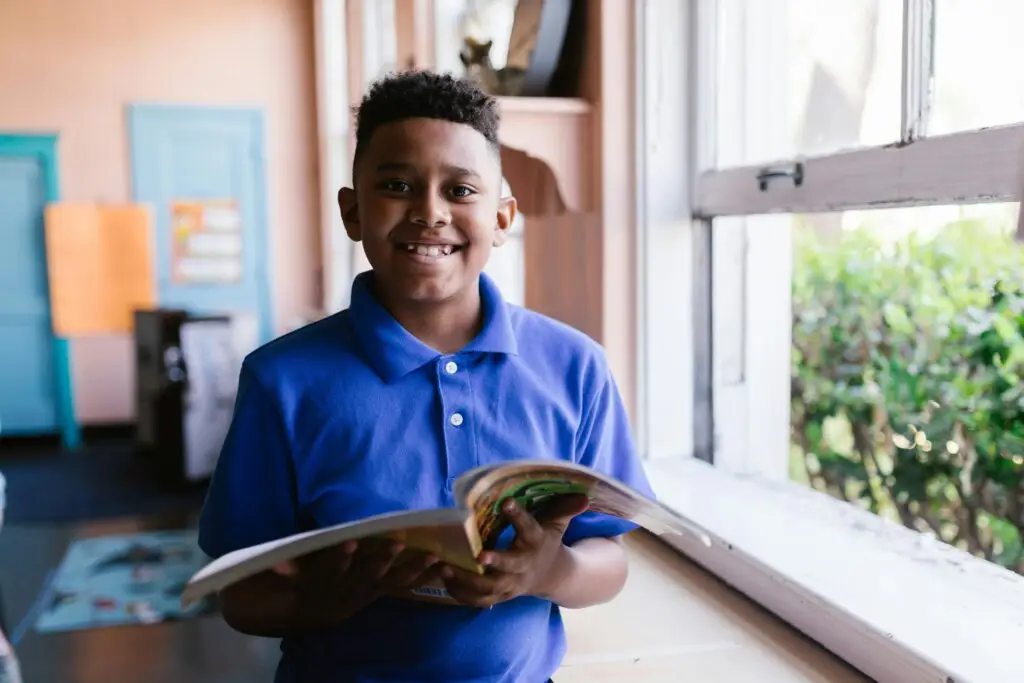 Young boy in blue polo reading by a classroom window, smiling happily.